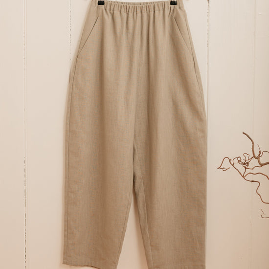 A pair of beige barrel leg trousers hanging on a hanger against a white wall, with a slim elasticated waistband and a high-waisted cut.