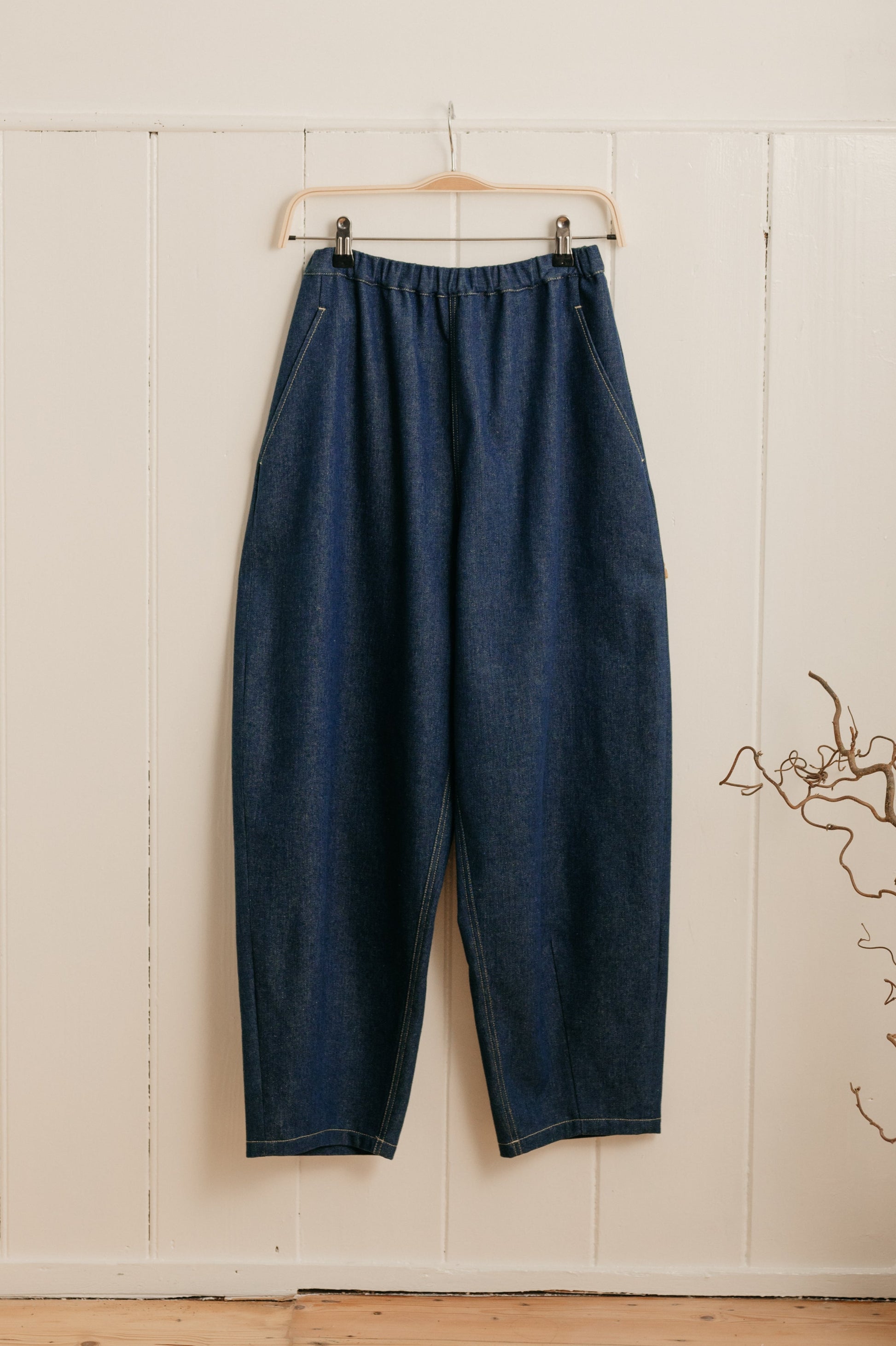 A pair of indigo denim barrel leg trousers with an elasticated waistband hanging on a hanger against a white wooden background.