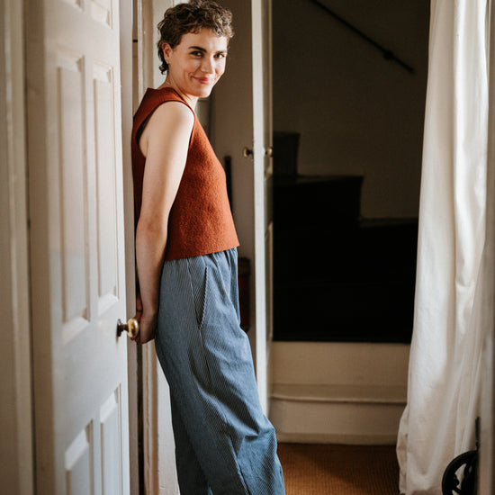 Woman standing in a doorway wearing a rust-coloured sleeveless top and blue striped denim trousers