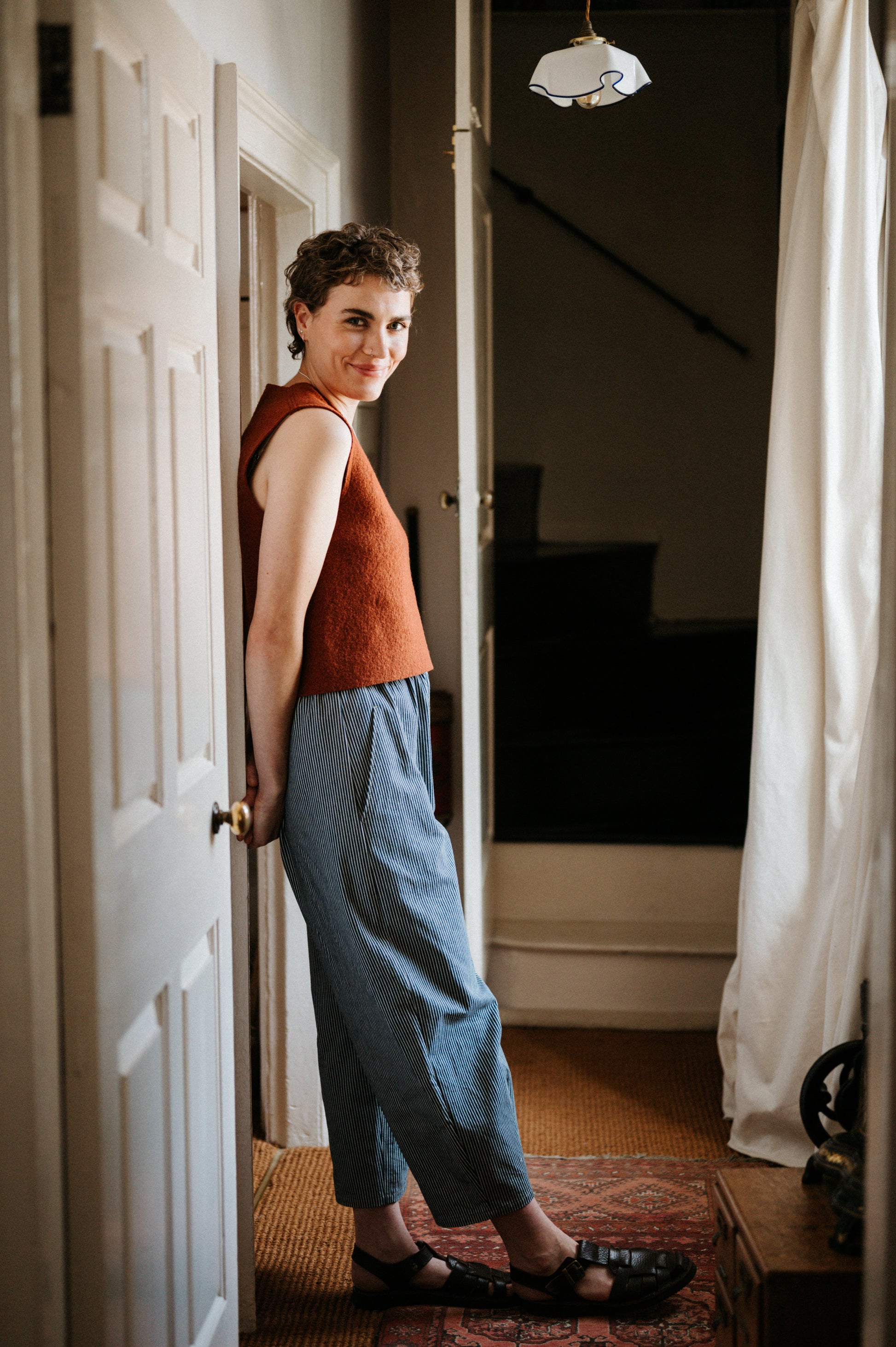 Woman standing in a doorway wearing a rust-coloured sleeveless top and blue striped denim trousers