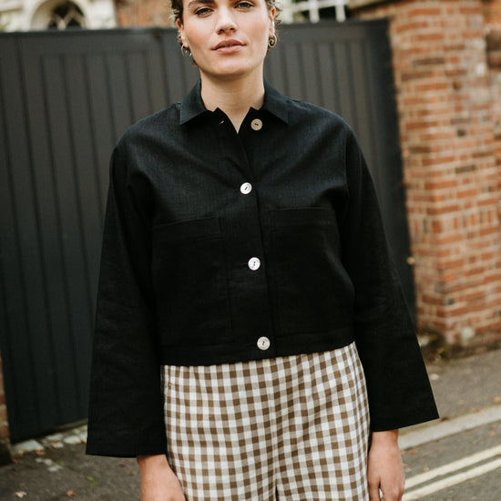 Person wearing a black jacket and checkered pants standing in front of a brick building.