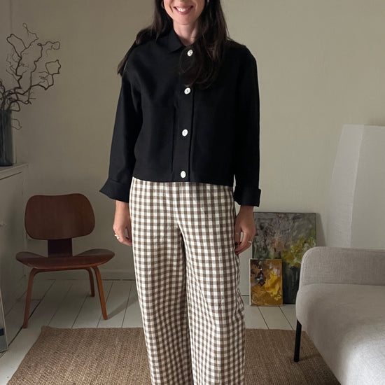 Woman standing in a living room wearing a black workwear jacket and brown gingham trousers.