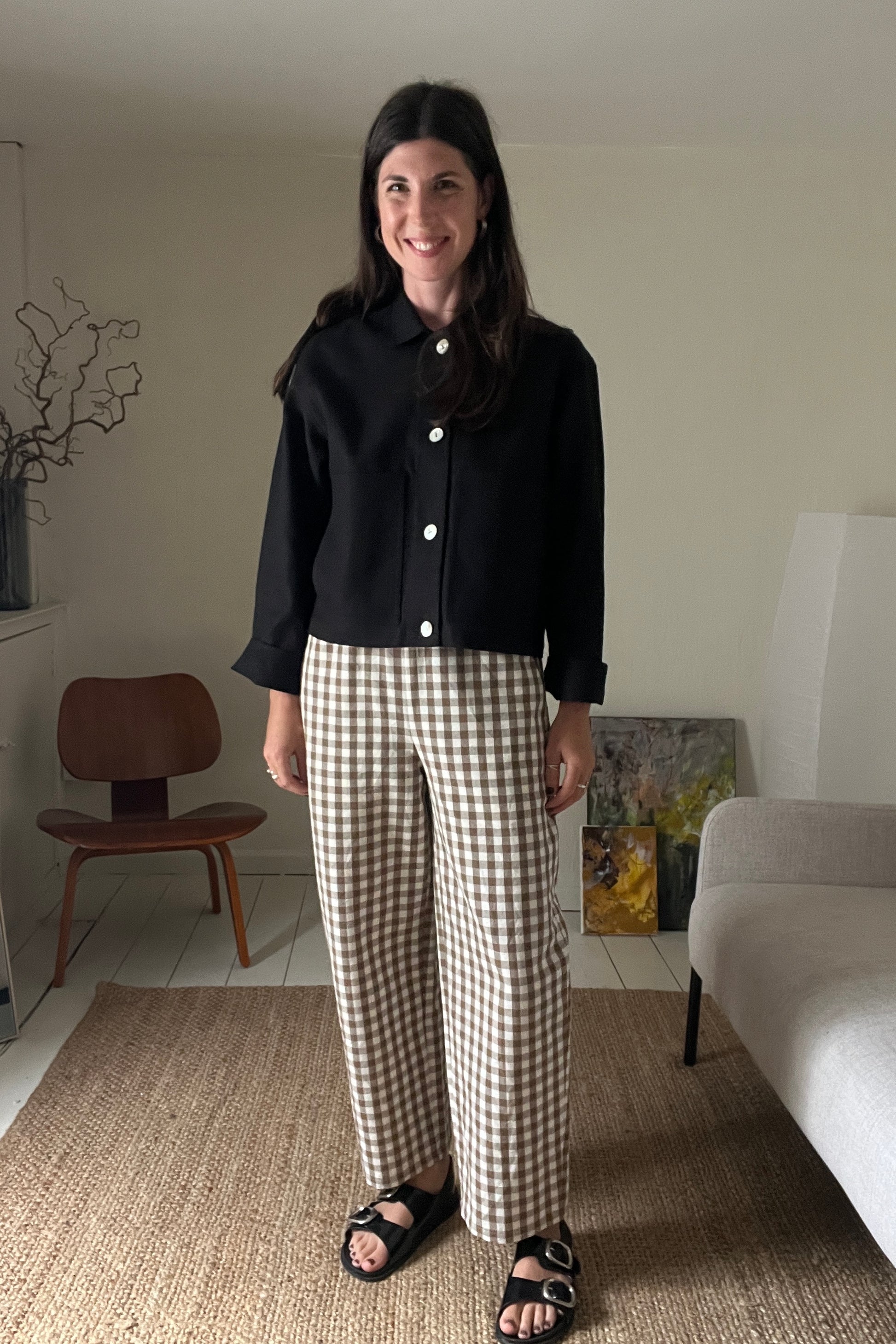 Woman standing in a living room wearing a black workwear jacket and brown gingham trousers.