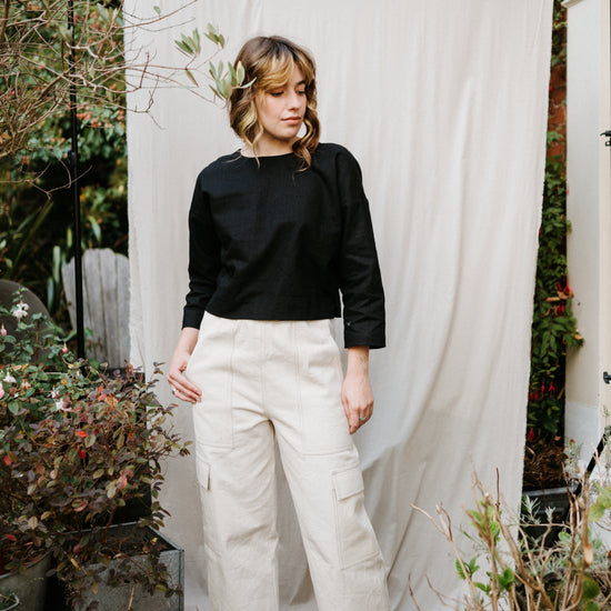 Person wearing a black top and ecru cargo jeans standing in front of a white curtain with plants around.