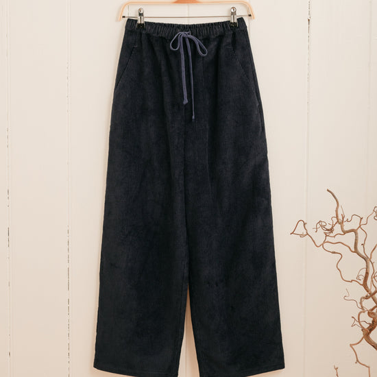 A pair of navy corduroy trousers with a drawstring waistband hanging on a hanger against a white background, with a subtle blue cord drawstring detail.