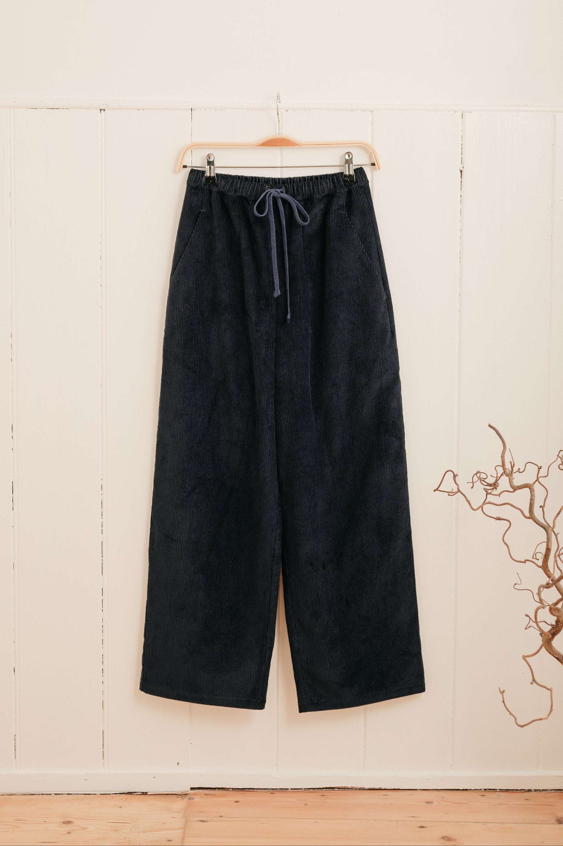 A pair of navy corduroy trousers with a drawstring waistband hanging on a hanger against a white background, with a subtle blue cord drawstring detail.