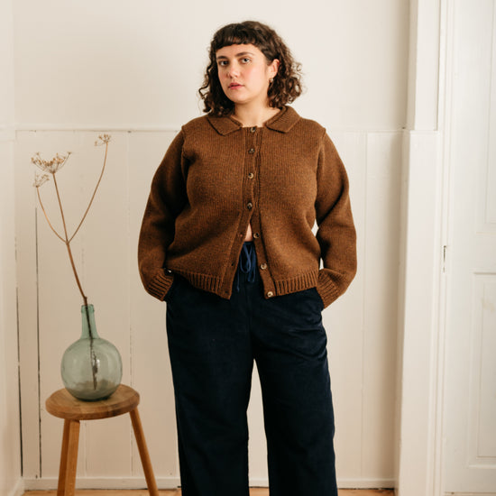 model wearing navy cord trousers and a brown cardigan standing next to a stool with a vase on