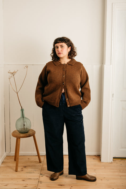 model wearing navy cord trousers and a brown cardigan standing next to a stool with a vase on