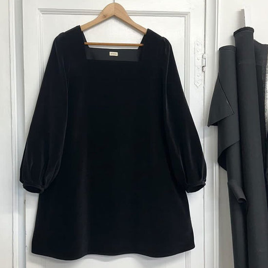 A black velvet mini dress with a square neckline and bell sleeves hanging on a hanger against a white door backdrop, with clothing items in the background to the right.