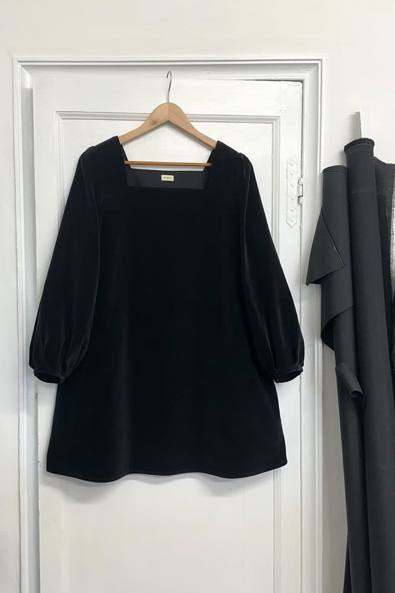 A black velvet mini dress with a square neckline and bell sleeves hanging on a hanger against a white door backdrop, with clothing items in the background to the right.