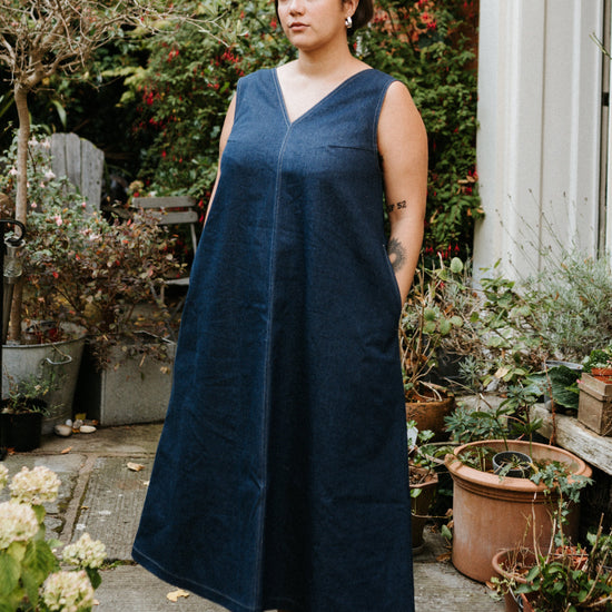 Woman in a blue denim dress standing in a garden with plants and a house in the background