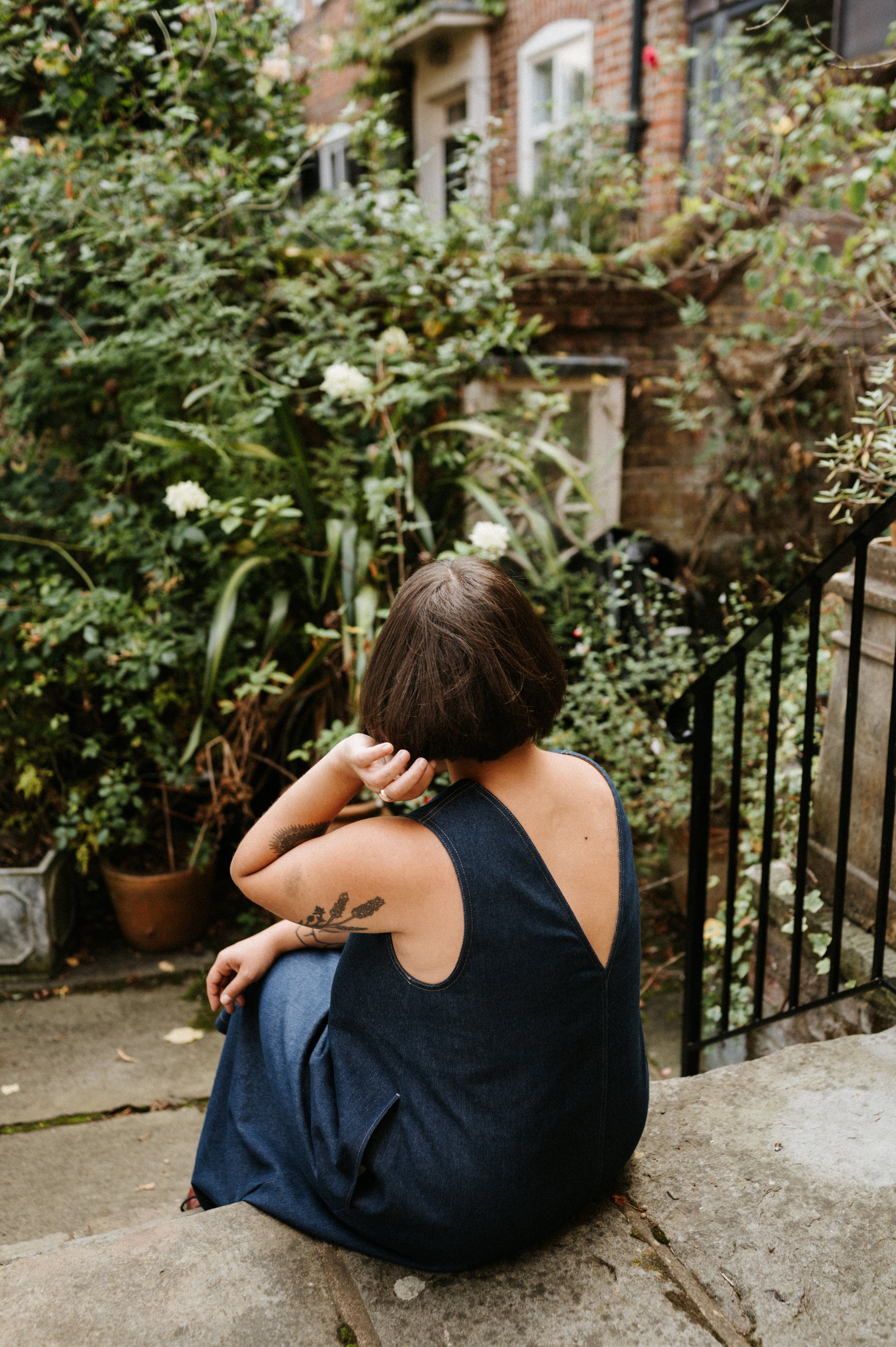 Person sitting on a stone wall in a garden with greenery and a house in the background