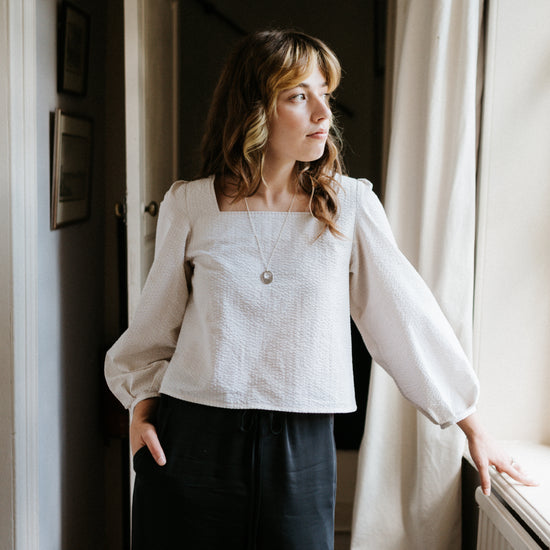 Woman standing by a window wearing a white blouse and dark pants in a softly lit room.