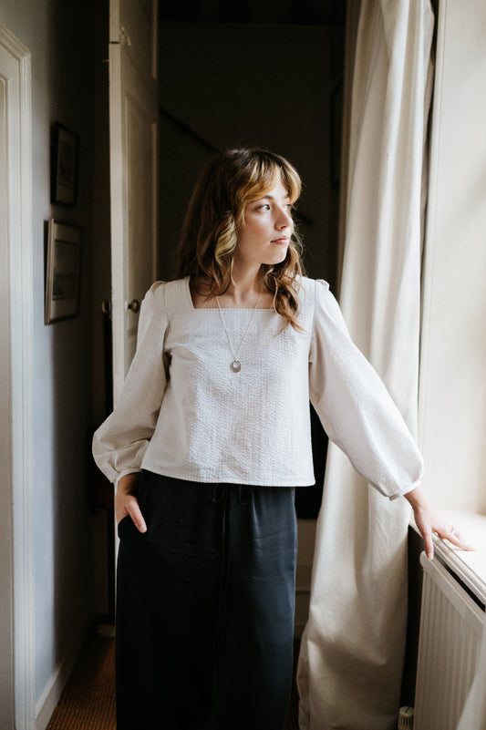 Woman standing by a window wearing a white blouse and dark pants in a softly lit room.