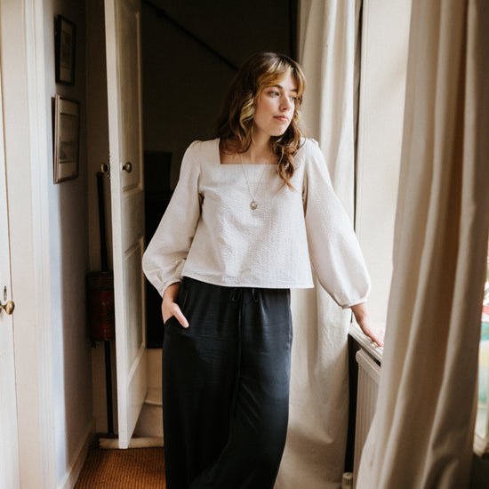 Woman in a white blouse and dark pants standing in a room with a window and curtain.