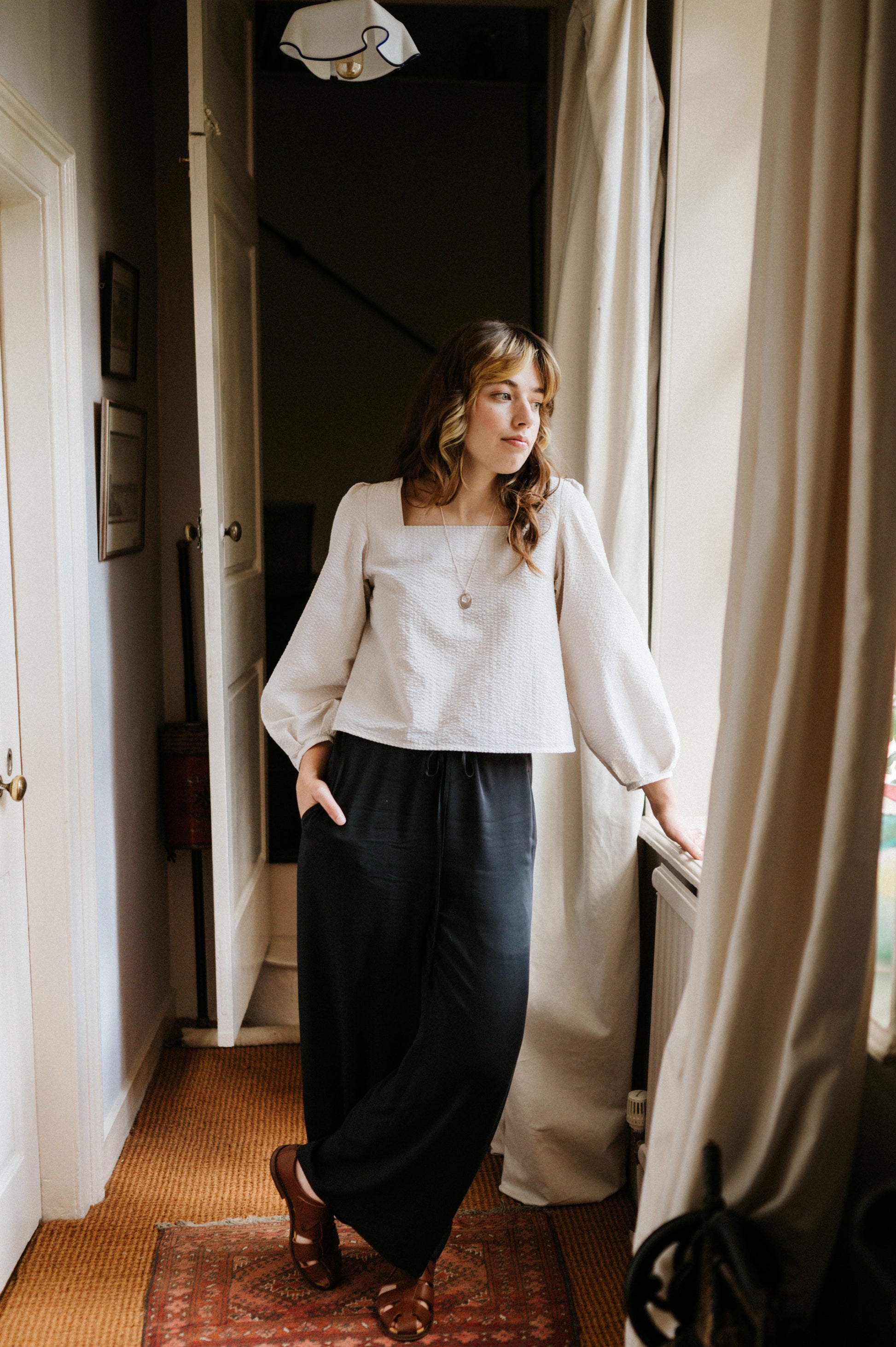 Woman in a white blouse and dark pants standing in a room with a window and curtain.