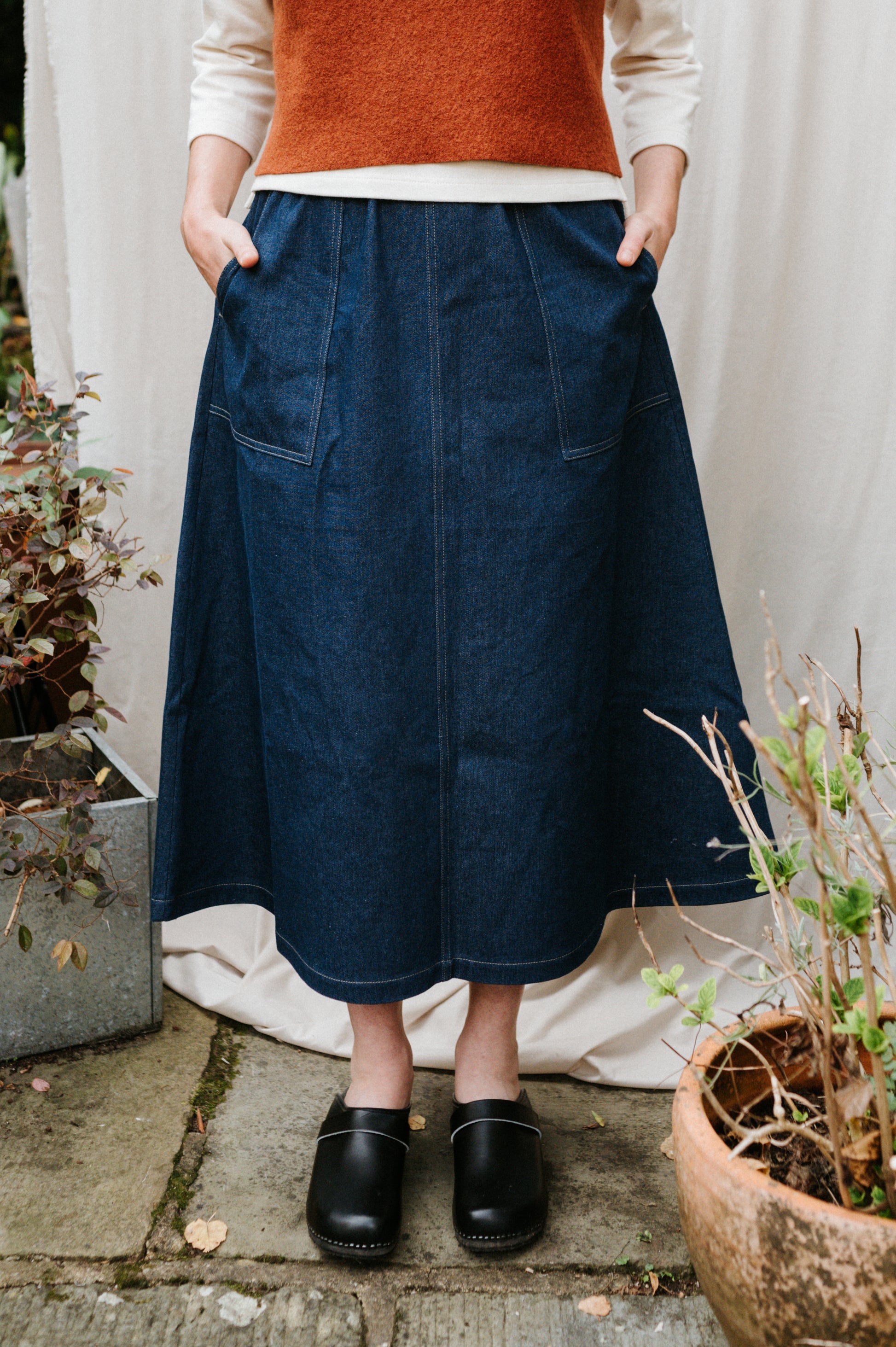 Person wearing a blue denim utility skirt with plants and a white curtain in the background