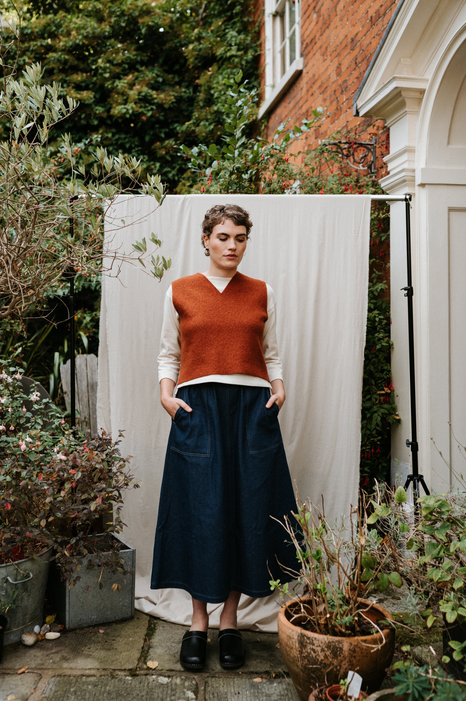 Person wearing a rust-colored sweater and dark blue skirt standing outdoors with plants and a white backdrop.