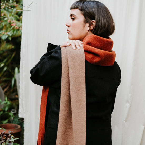 Person wearing a black coat with a brown scarf and orange sweater against a white curtain.