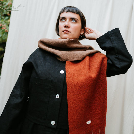 Person wearing a black coat and brown and rust scarf against a white curtain with greenery.