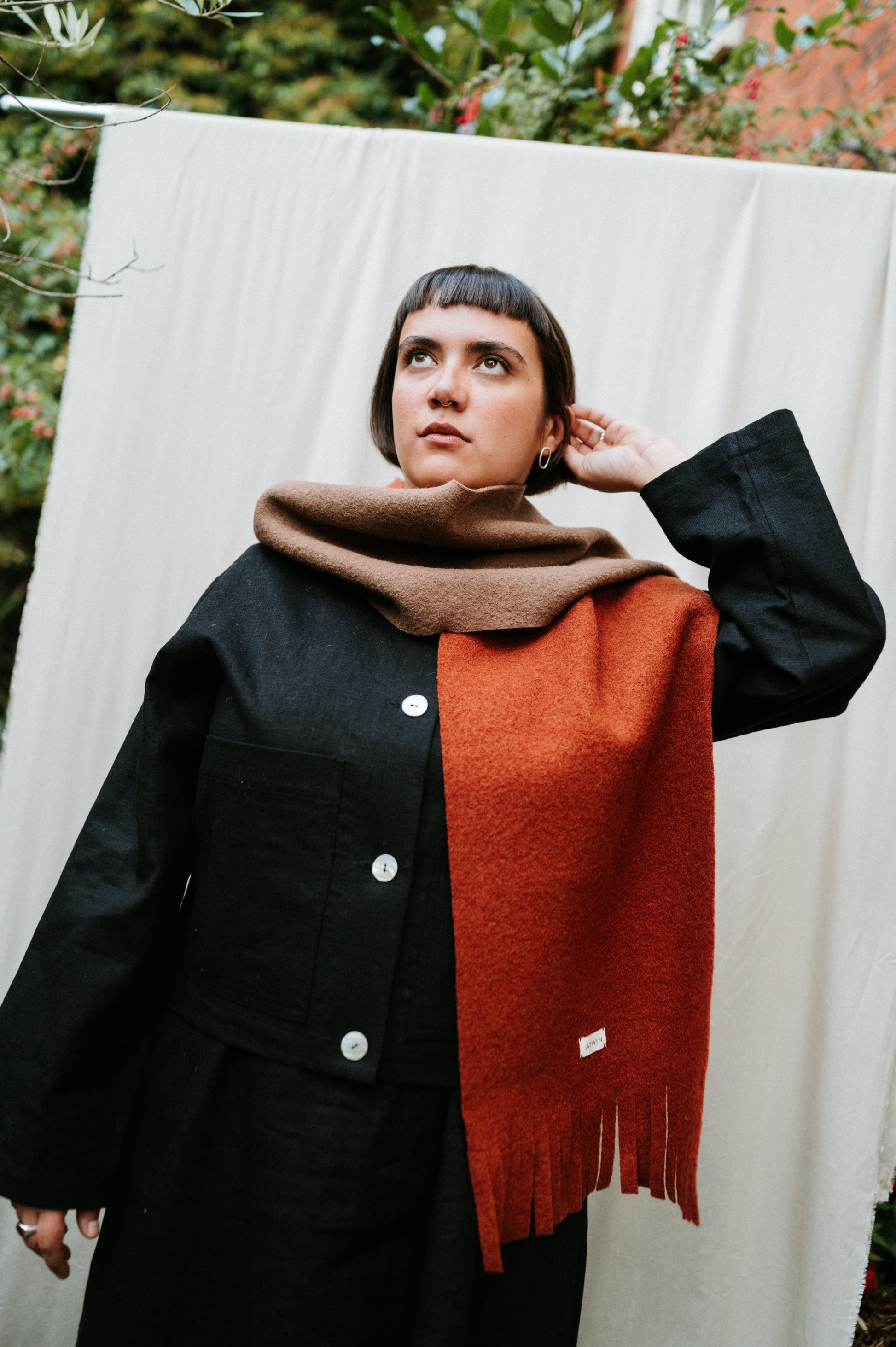 Person wearing a black coat and brown and rust scarf against a white curtain with greenery.