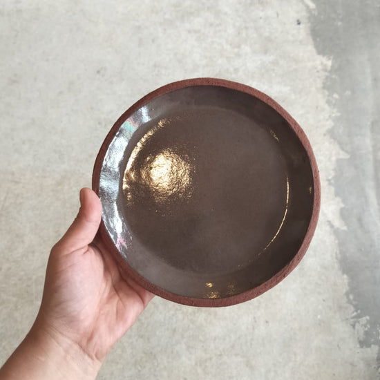 A hand holding a round terracotta ceramic side plate with a glossy finish.