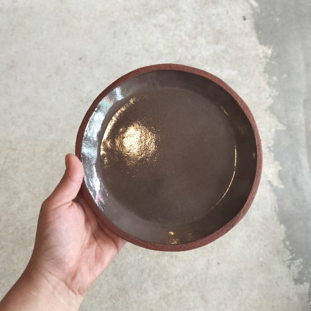 A hand holding a round terracotta ceramic side plate with a glossy finish.