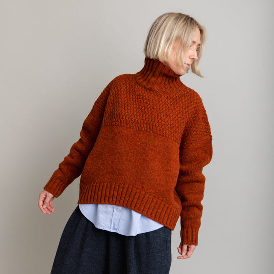 Person wearing a rust-colored knitted sweater and dark pants on a plain background