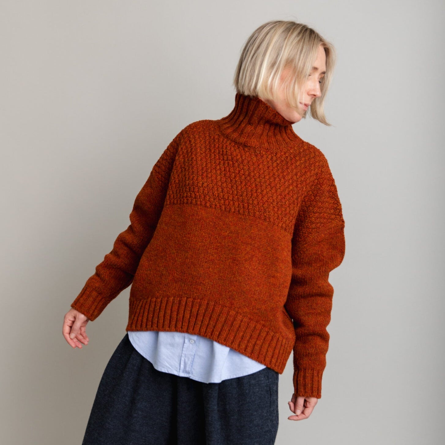 Person wearing a rust-colored knitted sweater and dark pants on a plain background