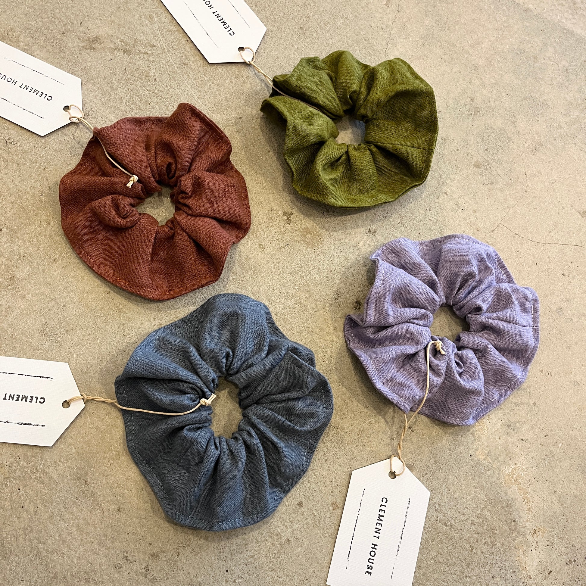 Four scrunchies in brown, green, blue, and purple on a textured surface with tags.