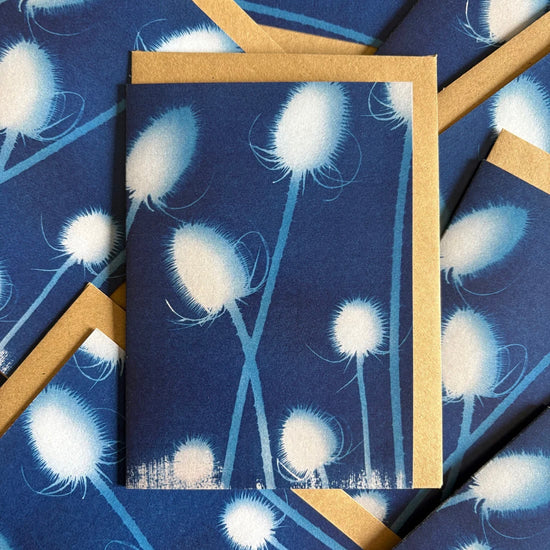 Greeting card with white floral teasel design on a blue background with a brown envelope tucked behind, surrounded by matching cards with envelopes