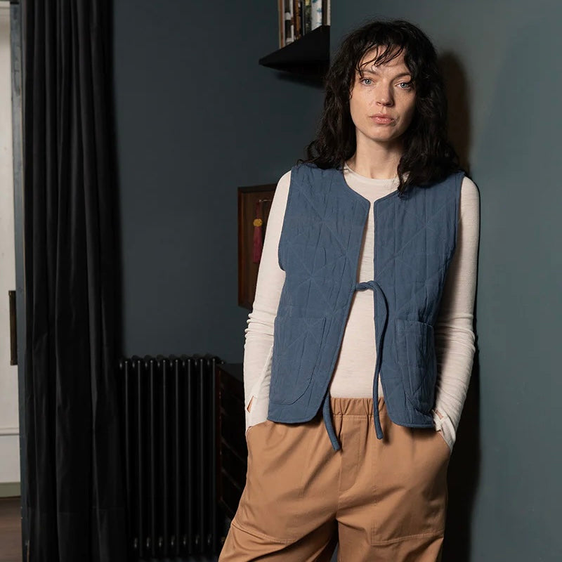 Female model with dark brown hair wears blue padded quilted gilet with a single front tie. The model has a white long sleeved crew neck top on underneath and camel coloured trousers standing against a blue wall in a home