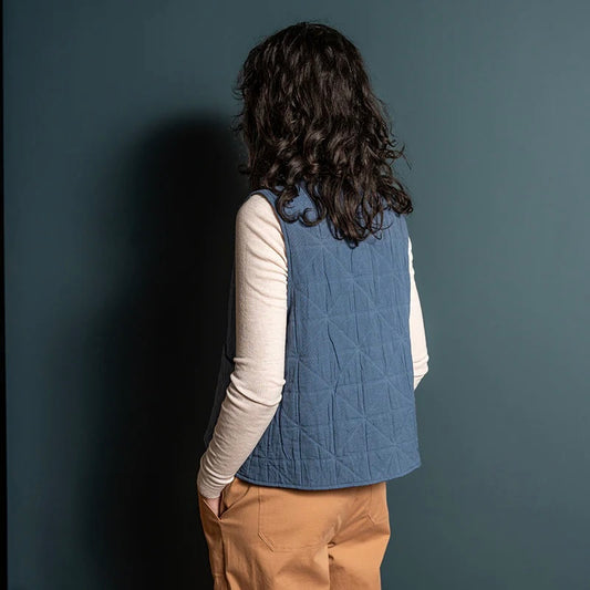 Female model with dark brown hair wears blue padded quilted gilet with a single front tie. The model has a white long sleeved crew neck top on underneath and camel coloured trousers standing against a blue wall in a home