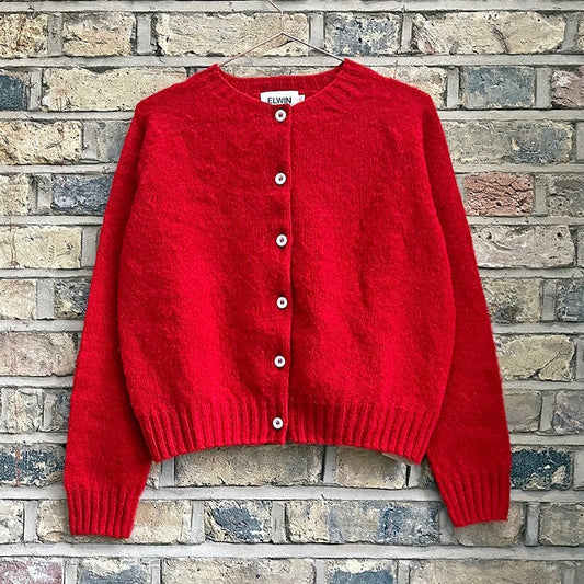A red knitted cardigan with buttons and ribbed hem laid flat on a brick surface.