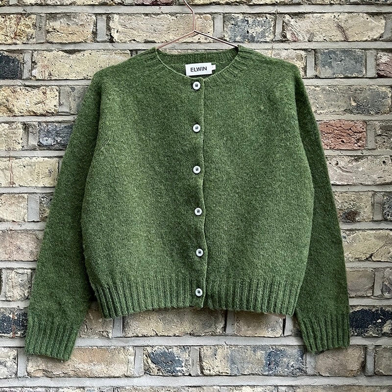 Green knitted cardigan with buttons and ribbed hem, displayed on a hanger against a brick wall.