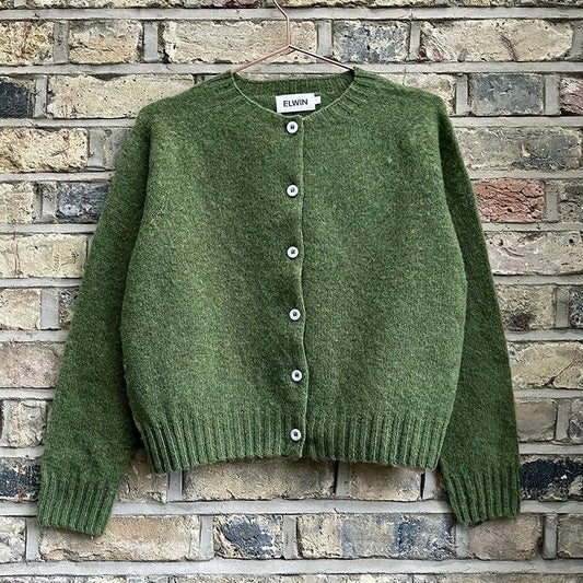 Green knitted cardigan with buttons and ribbed hem, displayed on a hanger against a brick wall.