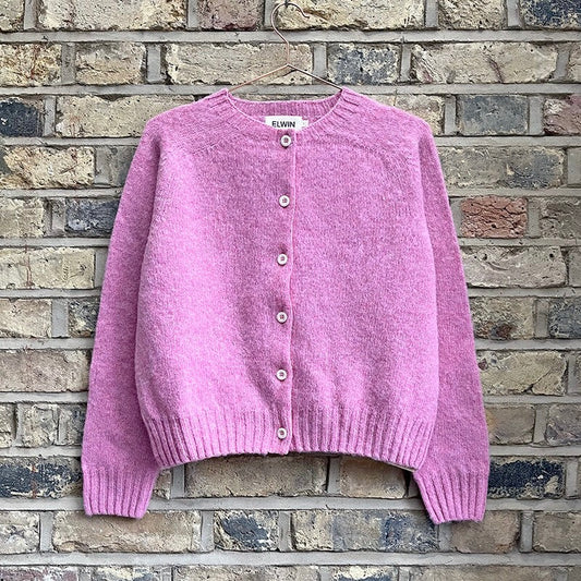 A pink knitted crew neck cardigan with button closures and rib knit cuffs displayed against a brick wall.