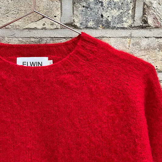 Red sweater with a brand label on a hanger against a brick wall.