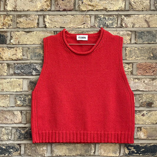 A flame orange knitted vest with a round neckline, displayed against a brick wall background.
