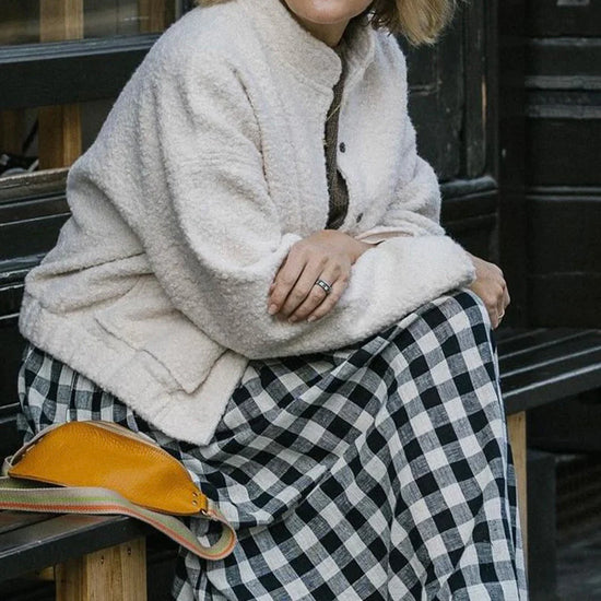 Woman sitting on a bench wearing a cream jacket and black and white checkered skirt.