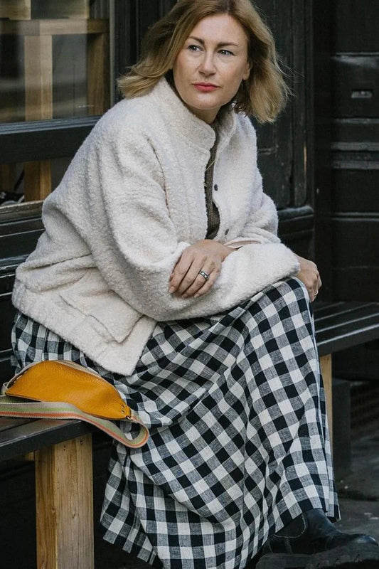 Woman sitting on a bench wearing a cream jacket and black and white checkered skirt.