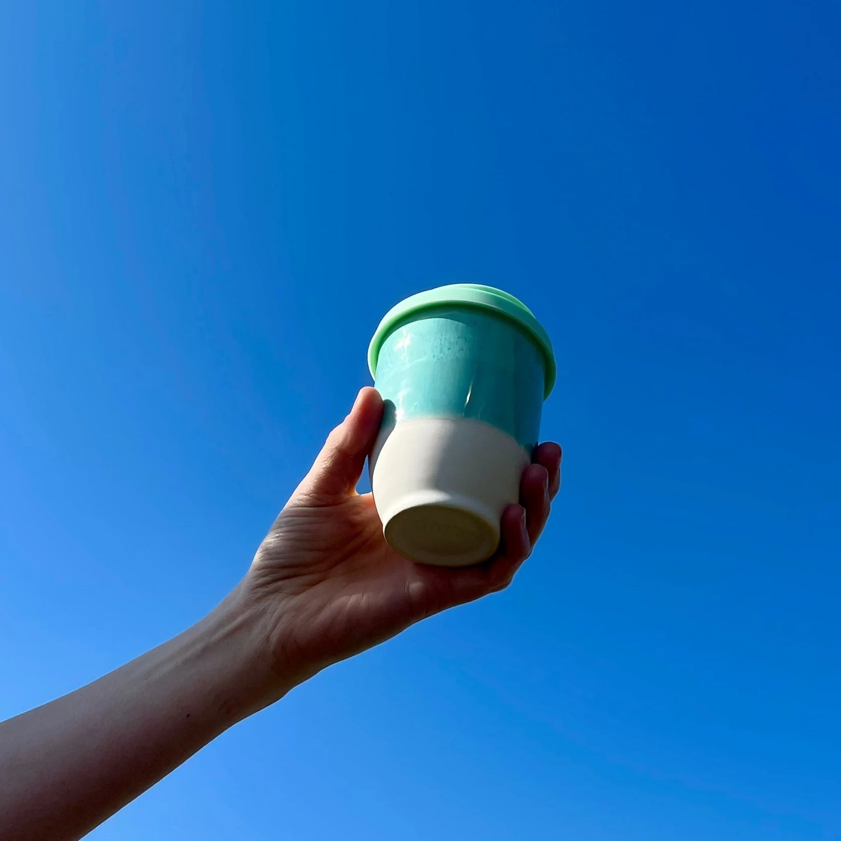 Hand holding a two-toned green and white cup against a clear blue sky