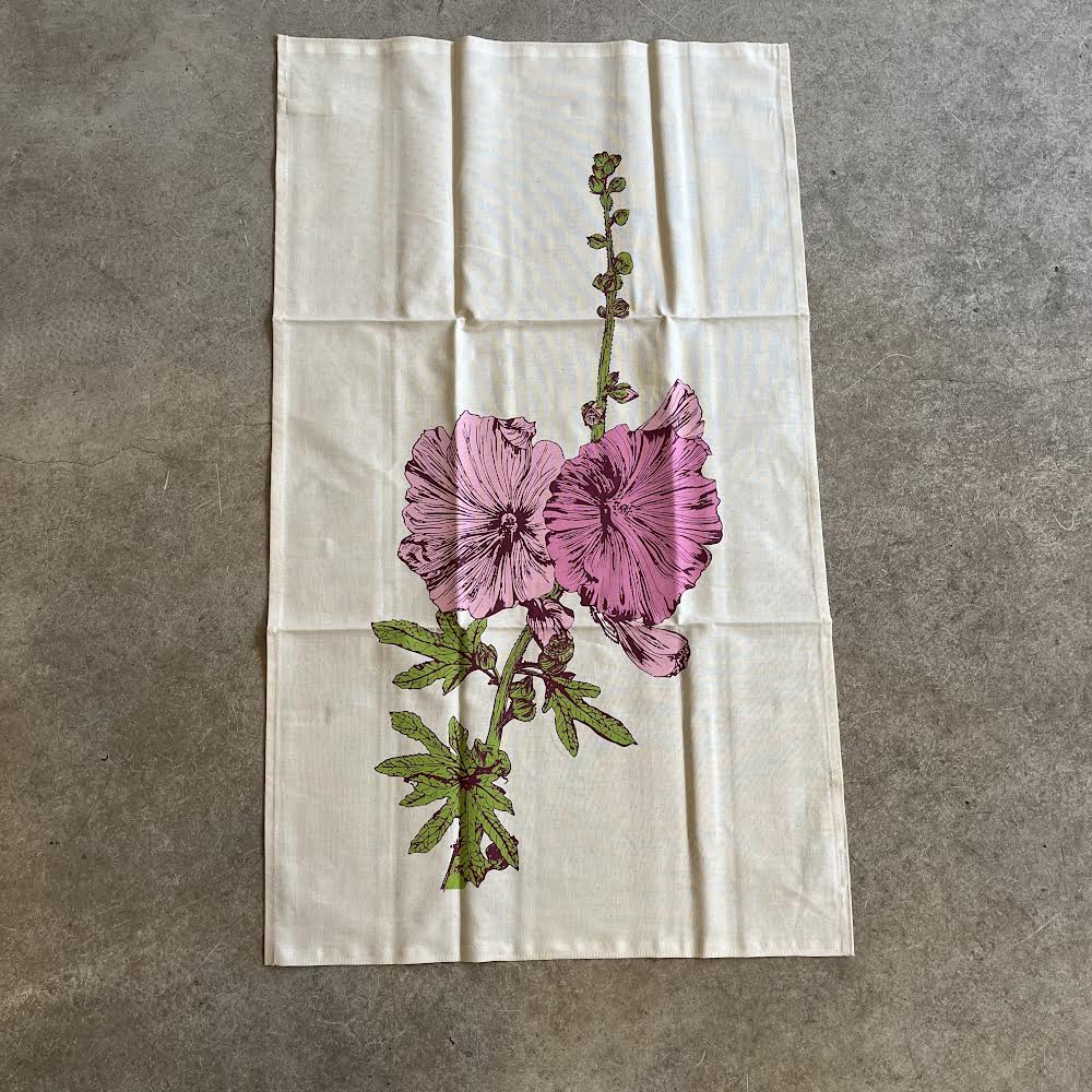 A beige cotton tea towel with a floral hollyhock design printed on it, laid out flat.
