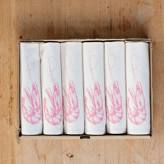A set of six folded cloth napkins with a pink prawn design, placed in a cardboard box on a wooden surface.