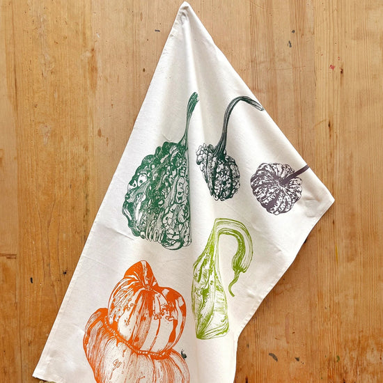 Tea towel with autumnal design of pumpkins and gourds hanging on a wooden wall with real pumpkins and gourds below.