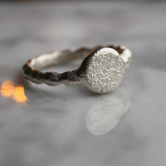A silver ring with a textured surface and a rounded profile, displayed on a reflective surface with a blurred background.