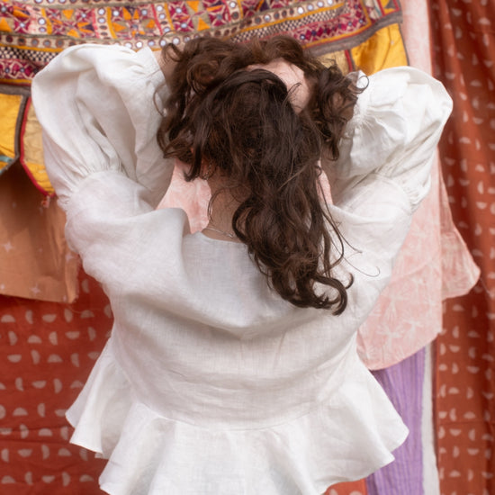 Person wearing a white blouse with ruffled details and green patterned pants, standing in front of colorful fabric.
