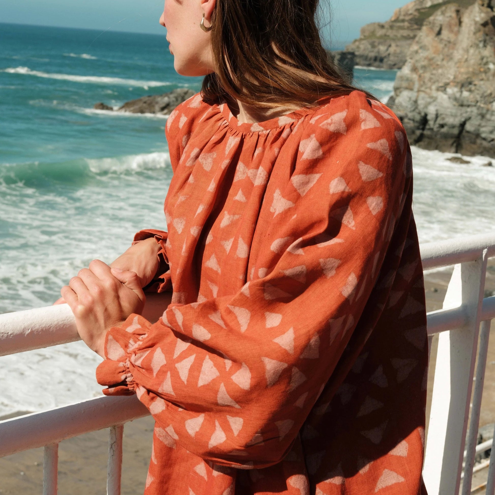 Woman in an orange dress with a geometric pattern standing by a railing overlooking the ocean.