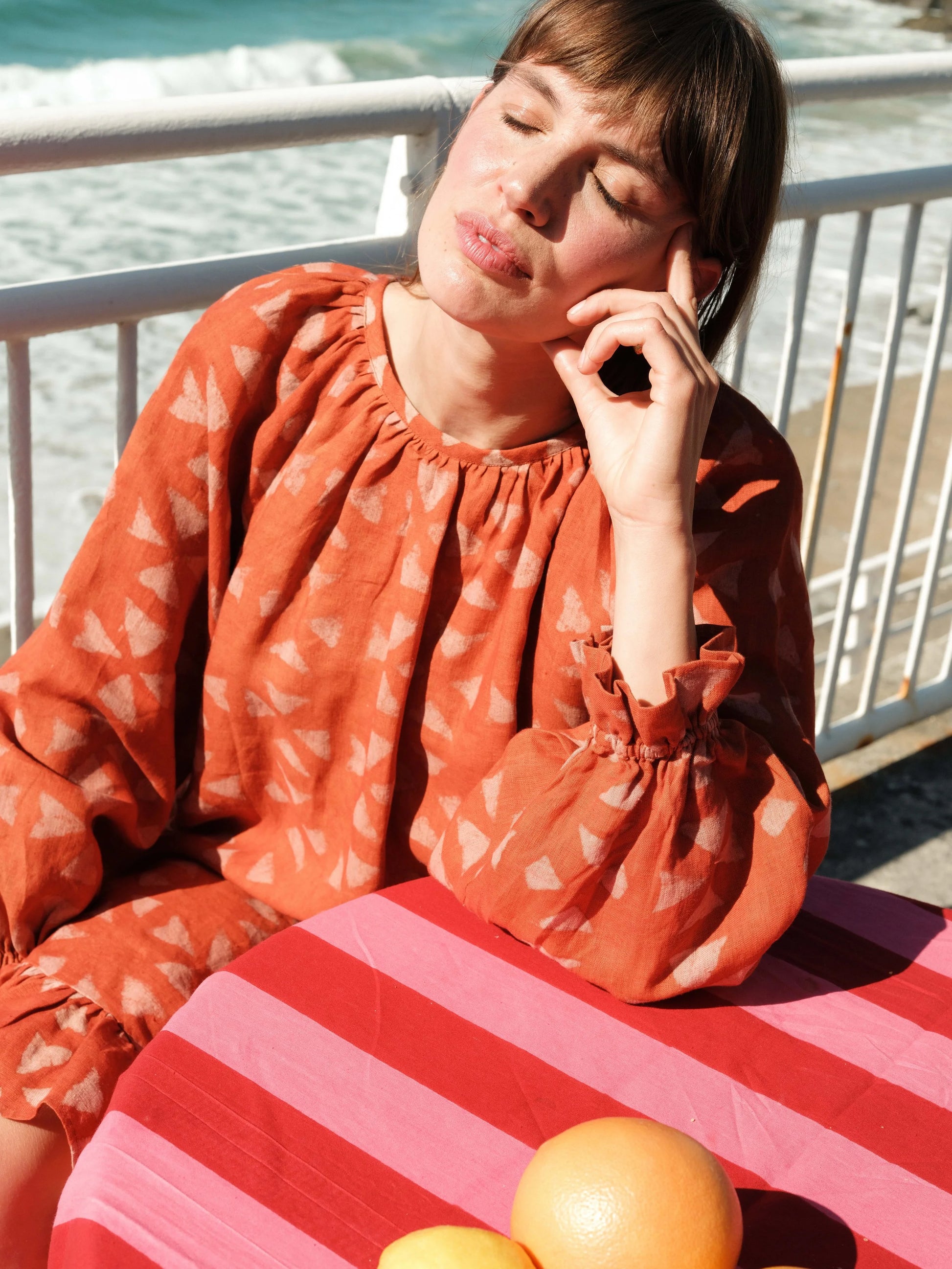 Woman in an orange dress sitting at a table with striped tablecloth by the sea.