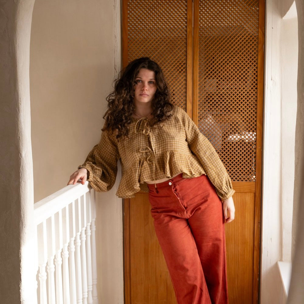A person wearing a flaxen-colored top with balloon sleeves and a front tie feature, paired with red pants, standing near a door with a wooden frame.